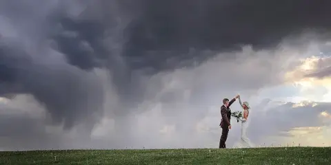 A bride and groom dancing in a grassy field under a dramatic, cloudy sky.