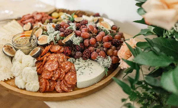 A large wooden platter filled with an assortment of cured meats, various cheeses, fresh figs, grapes, and crackers, set on a table next to a floral arrangement.
