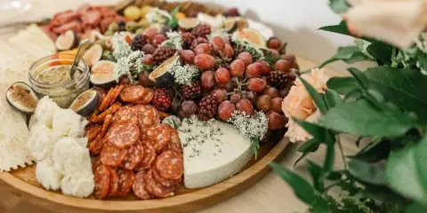 A large wooden platter filled with an assortment of cured meats, various cheeses, fresh figs, grapes, and crackers, set on a table next to a floral arrangement.