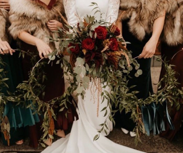 Bride in a white gown holding a large bouquet of red roses and greenery, flanked by bridesmaids holding a floral wreath.