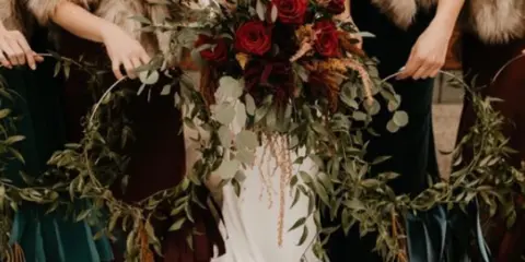Bride in a white gown holding a large bouquet of red roses and greenery, flanked by bridesmaids holding a floral wreath.
