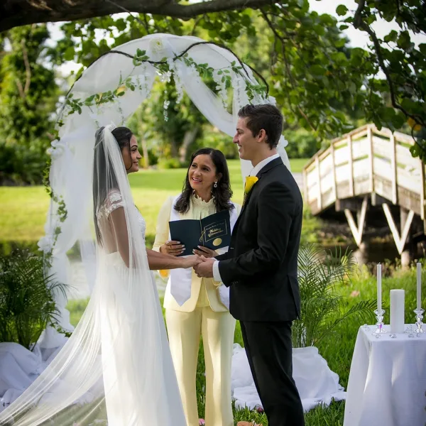 A bride and groom stand under a floral arch during an outdoor wedding ceremony, holding hands while an officiant in a cream suit stands between them.