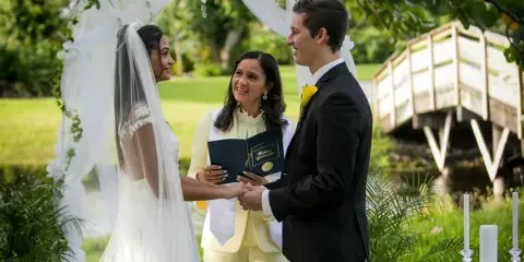 A bride and groom stand under a floral arch during an outdoor wedding ceremony, holding hands while an officiant in a cream suit stands between them.