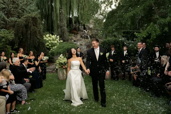 A bride and groom walk hand-in-hand down a grassy aisle lined with guests and floral arrangements, with petals falling around them.