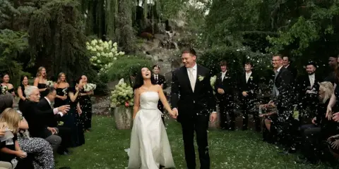 A bride and groom walk hand-in-hand down a grassy aisle lined with guests and floral arrangements, with petals falling around them.