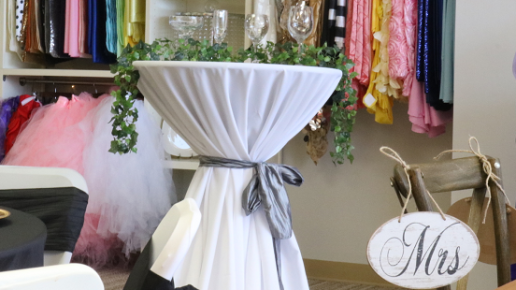 A white-draped cocktail table decorated with greenery and a 'Mrs' sign in a showroom setting.