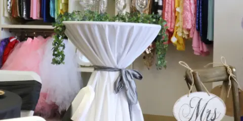 A white-draped cocktail table decorated with greenery and a 'Mrs' sign in a showroom setting.