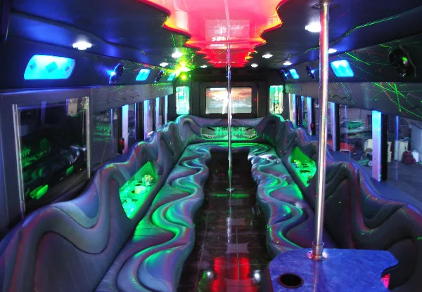 Interior of a party bus with a central dance pole, wet bar, and colorful party lighting.