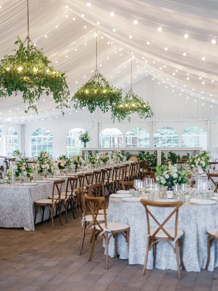 Elegant wedding reception interior with white tent ceiling, hanging greenery chandeliers, and floral table centerpieces.