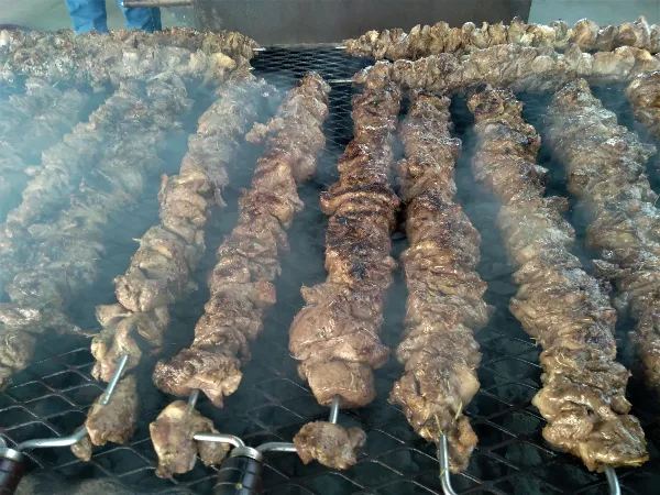 Rows of meat skewers grilling on a large outdoor barbecue grill with visible smoke.
