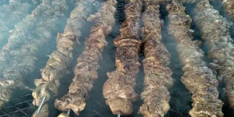 Rows of meat skewers grilling on a large outdoor barbecue grill with visible smoke.