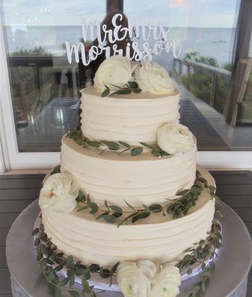 Three-tier white wedding cake with white roses and eucalyptus and a Mr & Mrs Morrison topper, displayed by a window with an ocean view.