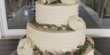 Three-tier white wedding cake with white roses and eucalyptus and a Mr & Mrs Morrison topper, displayed by a window with an ocean view.
