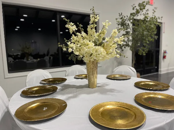 Interior view of an event venue showing a round table set with white tablecloth, gold charger plates, and a tall floral centerpiece.