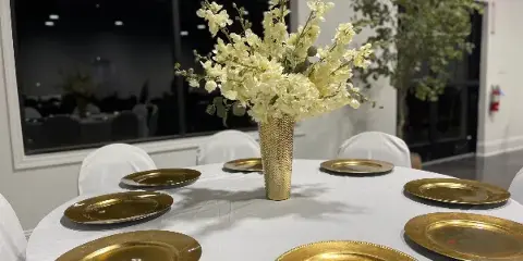 Interior view of an event venue showing a round table set with white tablecloth, gold charger plates, and a tall floral centerpiece.