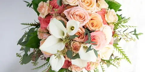A close-up of a wedding bouquet held by a bride, featuring peach roses, white calla lilies, and greenery.