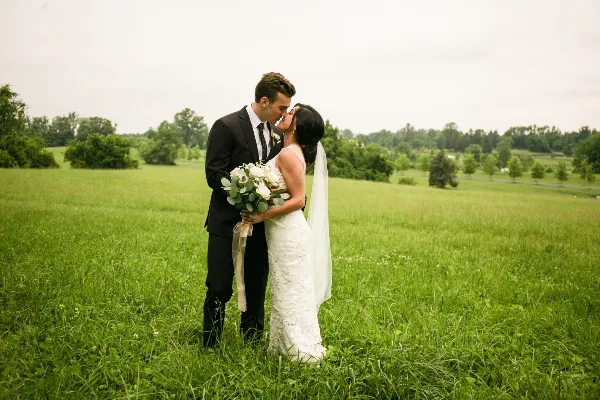 A bride and groom standing in a grassy field, the bride holding a white bouquet while they share a kiss.