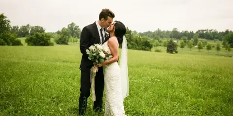 A bride and groom standing in a grassy field, the bride holding a white bouquet while they share a kiss.