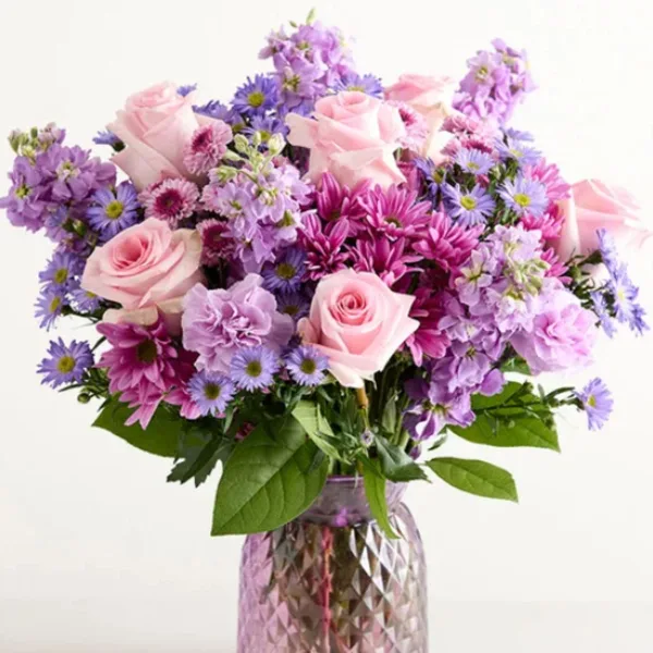 A floral arrangement featuring pink roses, purple daisies, and pink chrysanthemums in a clear glass vase.