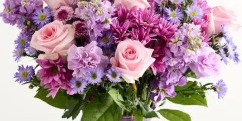 A floral arrangement featuring pink roses, purple daisies, and pink chrysanthemums in a clear glass vase.