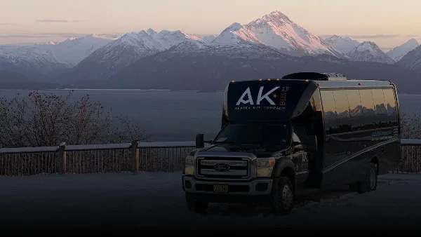 A black Alaska Bus Company shuttle bus parked in front of a scenic view of snow-capped mountains and a body of water.
