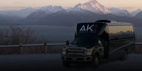 A black Alaska Bus Company shuttle bus parked in front of a scenic view of snow-capped mountains and a body of water.