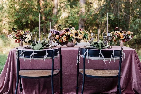 Styled outdoor wedding table with mauve linen, glassware, flatware, napkins, candles, and floral centerpieces.