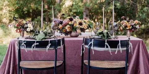 Styled outdoor wedding table with mauve linen, glassware, flatware, napkins, candles, and floral centerpieces.