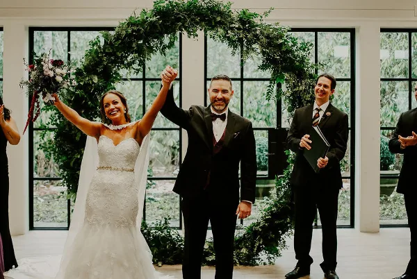 A bride and groom raise their joined hands in celebration during a wedding ceremony, with an officiant standing to the side.