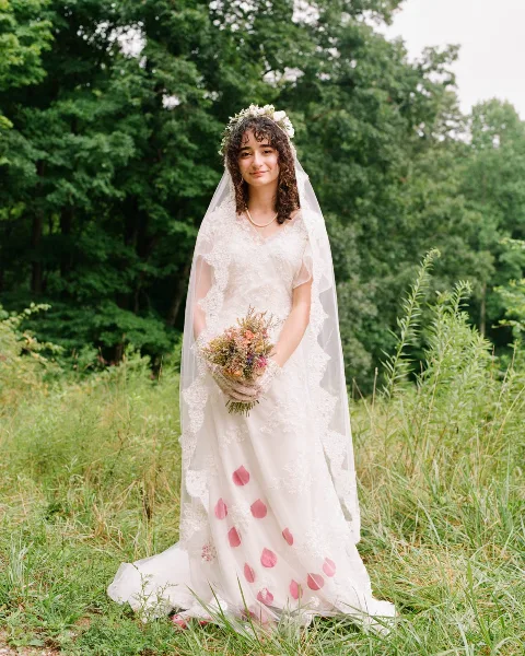 A bride wearing a white lace dress and veil stands in a grassy field holding a bouquet of flowers.