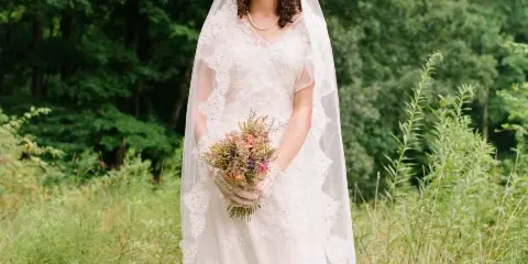 A bride wearing a white lace dress and veil stands in a grassy field holding a bouquet of flowers.