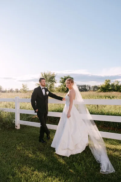 A bride and groom standing together in a field next to a white fence, captured in a professional wedding photograph.