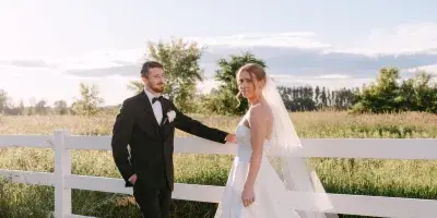 A bride and groom standing together in a field next to a white fence, captured in a professional wedding photograph.