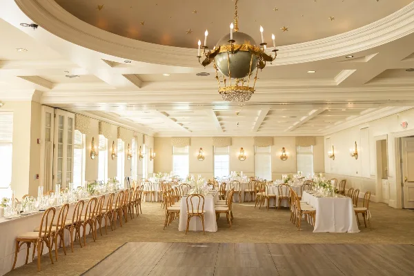 An elegant wedding reception hall featuring long banquet tables with white linens, wooden chairs, and a large ornate chandelier.