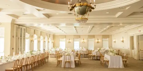 An elegant wedding reception hall featuring long banquet tables with white linens, wooden chairs, and a large ornate chandelier.