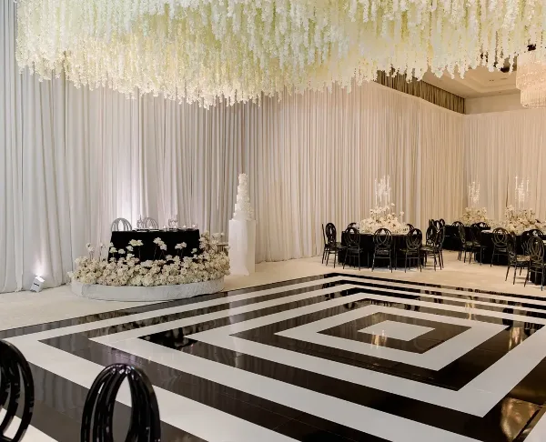 A wedding reception venue featuring a black and white geometric dancefloor, a large hanging floral installation, and decorated tables with black chairs.