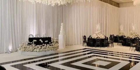 A wedding reception venue featuring a black and white geometric dancefloor, a large hanging floral installation, and decorated tables with black chairs.
