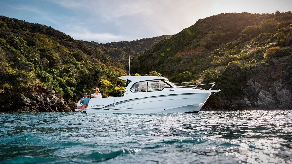 A white Beneteau Antares 8 motor yacht on calm water near a green hillside, with two people aboard, captured in a cinematic video still.