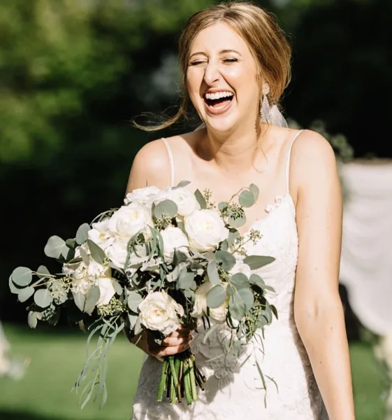A smiling bride in a white wedding dress holding a bouquet of white roses and eucalyptus, showcasing styled hair and makeup.