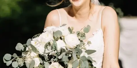 A smiling bride in a white wedding dress holding a bouquet of white roses and eucalyptus, showcasing styled hair and makeup.