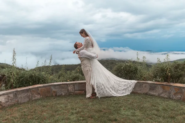 A bride in a long lace gown and veil is lifted by her groom in a white suit on a grassy hilltop with a stone wall and a cloudy sky in the background.