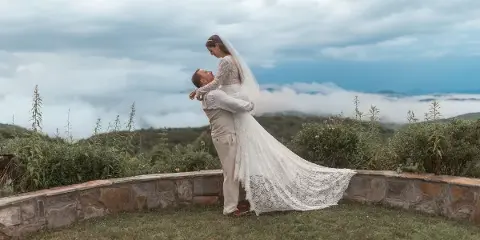 A bride in a long lace gown and veil is lifted by her groom in a white suit on a grassy hilltop with a stone wall and a cloudy sky in the background.