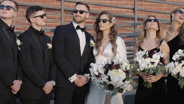 A bride and groom standing with their wedding party in formal attire, holding bouquets.