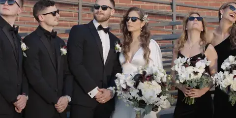 A bride and groom standing with their wedding party in formal attire, holding bouquets.
