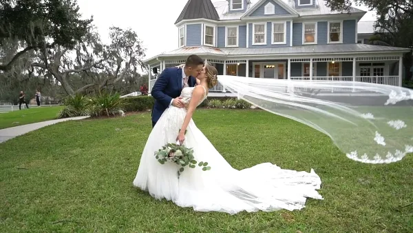 A bride and groom kissing on a lawn in front of a large blue house, with the bride's long veil flowing in the wind.