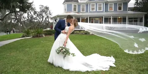 A bride and groom kissing on a lawn in front of a large blue house, with the bride's long veil flowing in the wind.