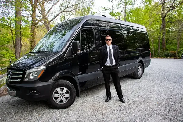 A professional driver in a black suit standing next to a black passenger van parked on a gravel driveway.