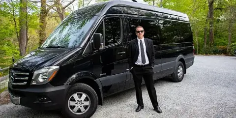 A professional driver in a black suit standing next to a black passenger van parked on a gravel driveway.