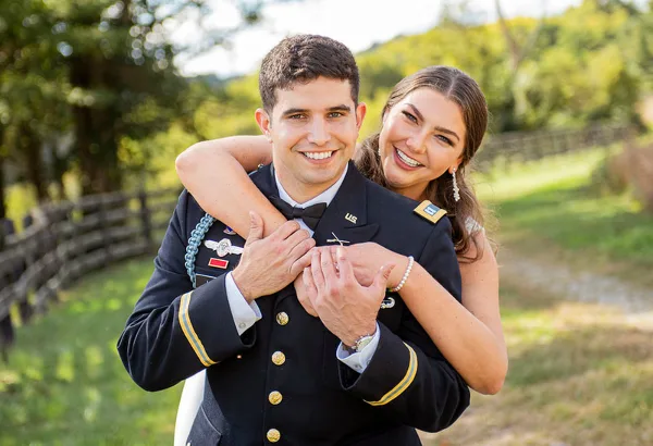 A smiling couple posing for a portrait outdoors, with the man wearing a military dress uniform and the woman in formal attire.