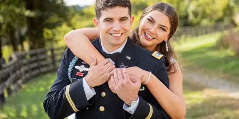 A smiling couple posing for a portrait outdoors, with the man wearing a military dress uniform and the woman in formal attire.